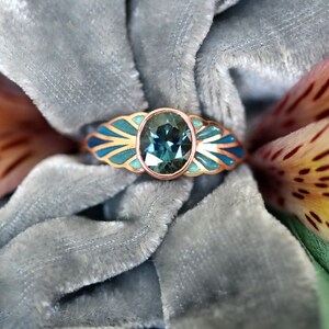 Puede incluir: Un anillo de oro rosa con una piedra ovalada azul engastada en un delicado dise&ntilde;o de inspiraci&oacute;n art d&eacute;co. El anillo est&aacute; rodeado por un dise&ntilde;o de esmalte azul y verde.