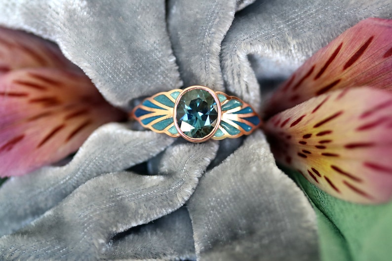 Puede incluir: Un anillo de oro rosa con una piedra ovalada azul engastada en un delicado dise&ntilde;o de inspiraci&oacute;n art d&eacute;co. El anillo est&aacute; rodeado por un dise&ntilde;o de esmalte azul y verde.