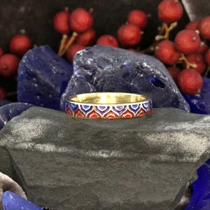 Puede incluir: Un anillo de banda de oro con un diseño de esmalte rojo y azul. El anillo está colocado sobre un fondo de piedras azules y grises.