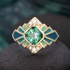 Puede incluir: Un anillo de oro con una piedra preciosa verde claro cuadrada en el centro. El anillo presenta un diseño geométrico con detalles de esmalte verde claro y azul verdoso y pequeños diamantes. El anillo está sobre una superficie oscura y texturizada.
