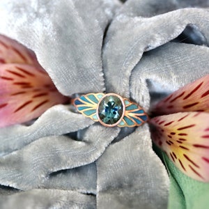 Puede incluir: Un anillo con una piedra preciosa azul engastada en una banda de oro. La banda est&aacute; decorada con esmalte azul y dorado. El anillo est&aacute; sobre una superficie de terciopelo gris.