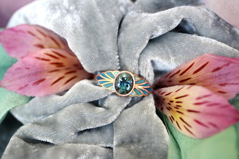 Puede incluir: Un anillo con una piedra preciosa azul engastada en una banda de oro. La banda est&aacute; decorada con esmalte azul y dorado. El anillo est&aacute; sobre una superficie de terciopelo gris.