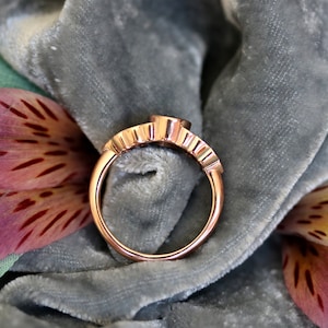 Puede incluir: Un anillo de oro rosa con una banda texturizada y una sola piedra redonda. El anillo se muestra sobre una superficie de terciopelo gris.