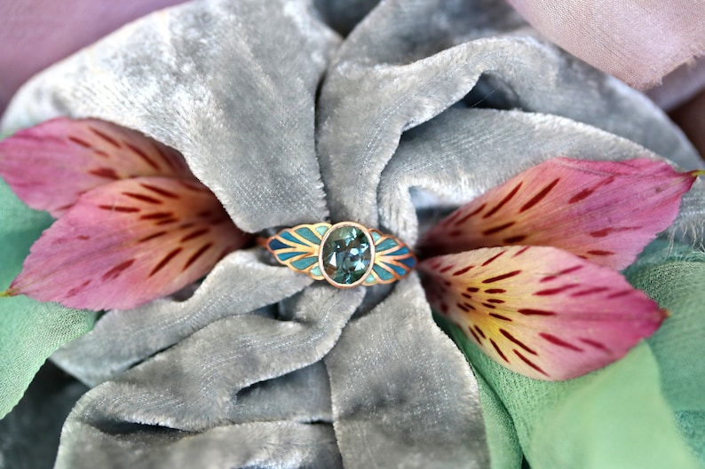 Puede incluir: Un anillo con una piedra ovalada azul engastada en una banda de oro. La banda est&aacute; decorada con esmalte azul y dorado, creando un dise&ntilde;o de hoja. El anillo se muestra sobre un fondo de terciopelo gris con p&eacute;talos de flores rosas.