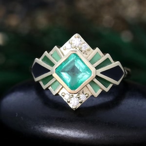 Puede incluir: Un anillo geométrico de esmeralda y diamante con detalles de esmalte negro. El anillo presenta una gran esmeralda talla esmeralda rodeada de diamantes más pequeños y esmalte negro.