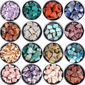 May include: A collection of sixteen circular metal bowls filled with various raw, rough-cut gemstones in shades of purple, green, red, blue, brown, white, orange, and pink, arranged in a four-by-four grid against a plain white background.