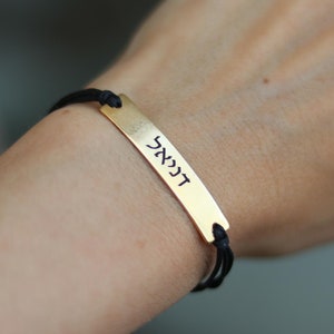 Puede incluir: Un brazalete de cordón negro con un dije de barra dorada grabado con la palabra hebrea "דבר".