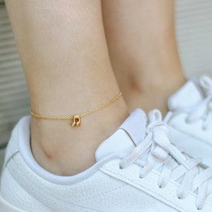 Puede incluir: Un brazalete de tobillo dorado con un pequeño dije en forma de nota musical. El brazalete se lleva en el tobillo de una persona, que lleva zapatillas blancas.