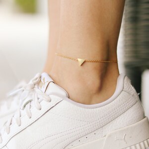 May include: A gold triangle anklet with a delicate chain. The anklet is worn on a person's ankle, with a white sneaker visible.