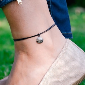 以下が含まれることがあります： 小さな貝殻のチャームが付いた黒いアンクレット。アンクレットは足首に着用され、青いジーンズとベージュのフラットシューズと組み合わせています。背景は緑の草です。