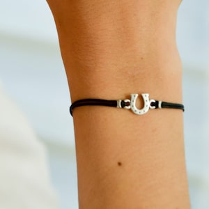 Puede incluir: Una pulsera de cuerda negra con un dije de herradura de plata. La pulsera está en la muñeca.