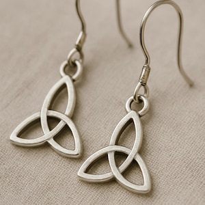 May include: A pair of silver-colored Celtic knot earrings. Each earring features a stylized triquetra design, suspended from a simple hook. The earrings are displayed against a neutral, textured background.