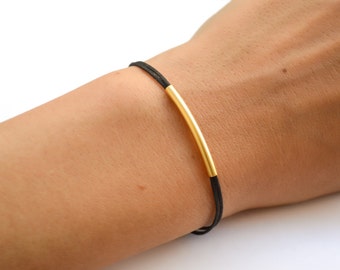 May include: A bracelet featuring a thin black cord and a single, smooth, curved gold-toned metal tube centered on the cord. The design consists of two strands of black cord that pass through the hollow gold-toned metal tube, creating a simple, sleek, and understated piece of jewelry.