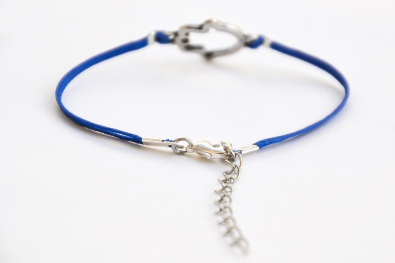 Pu&ograve; includere: Un braccialetto con cordino blu con un ciondolo e una chiusura a forma di cuore color argento. Il braccialetto ha un'estensione a catena per la regolazione delle dimensioni. Il gioiello &egrave; posto su uno sfondo bianco.