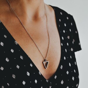 May include: A silver necklace with a geometric triangle pendant. The necklace is worn by a person wearing a black and white polka dot dress.