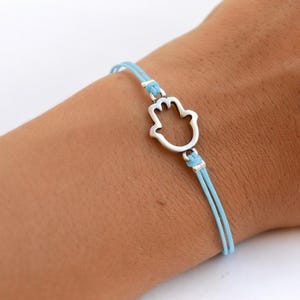 Puede incluir: Una pulsera de cordón azul claro con un dije de metal plateado en forma de mano de Hamsa. La pulsera está construida con dos cordones finos y paralelos de color azul claro que se conectan al dije central mediante pequeñas anillas plateadas.