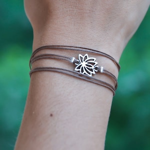 Puede incluir: Un brazalete de cordón marrón con un dije de loto plateado.