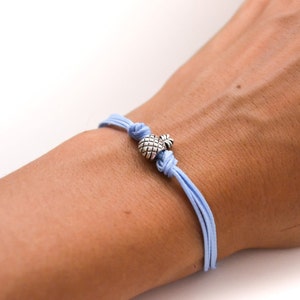 Puede incluir: Una pulsera trenzada azul claro con un dije de piña de plata.