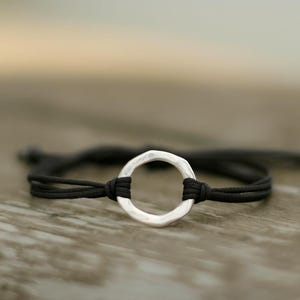 Op de afbeelding: Een minimalistische armband met een gehamerde zilverkleurige cirkel hanger aan een zwart koord. De armband wordt getoond op een houten oppervlak, wat het eenvoudige ontwerp benadrukt. De sieraden zijn geschikt voor dagelijks gebruik.