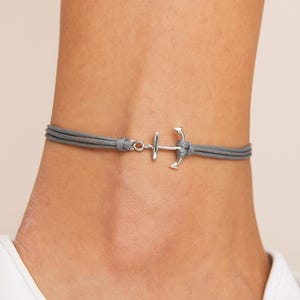 Peut inclure: Bracelet de cheville gris avec un charm en forme d'ancre argentée. Le bracelet est composé de trois brins de cordon gris, noués à chaque extrémité. Le charm en forme d'ancre est de style nautique.