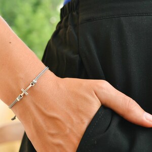 May include: A gray cord bracelet with a silver cross charm. The bracelet is worn on a person's wrist.