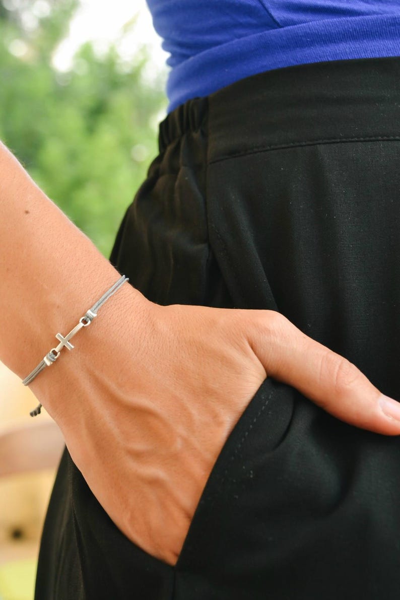 May include: A gray cord bracelet with a silver cross charm. The bracelet is worn on a person's wrist.