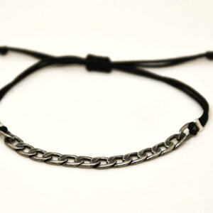 Pode incluir: Uma pulseira com um segmento de corrente de metal em tom prateado escuro ligado a um cordão preto. O cordão inclui um mecanismo de nó deslizante ajustável, permitindo modificar o comprimento para um ajuste seguro à volta do pulso.