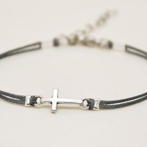 May include: A grey cord bracelet with a silver cross charm. The bracelet has a sliding clasp for adjusting the fit.