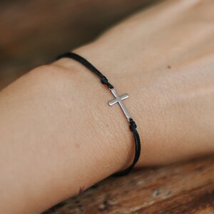 Puede incluir: Un brazalete de cuerda negra con un dije de cruz plateada.