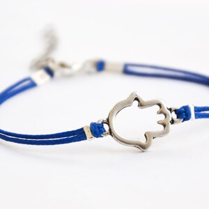 Pu&ograve; includere: Un braccialetto con cordino blu con un ciondolo a forma di mano di Hamsa color argento. Il braccialetto presenta un design a doppio cordino con dettagli bianchi e una chiusura a moschettone. La mano di Hamsa &egrave; un simbolo di protezione.