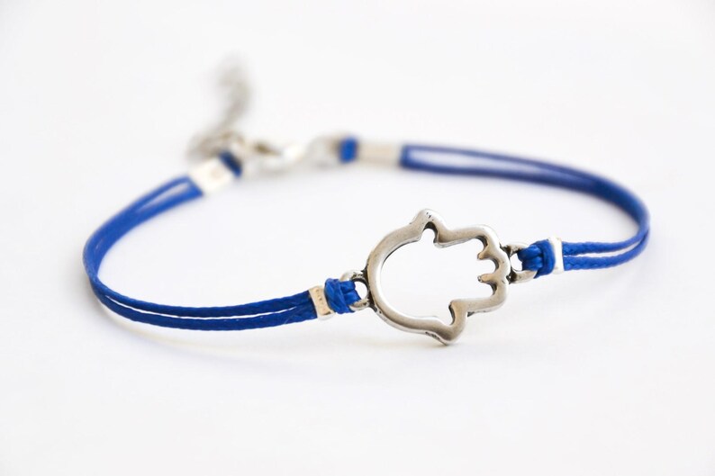 Pu&ograve; includere: Un braccialetto con cordino blu con un ciondolo a forma di mano di Hamsa color argento. Il braccialetto presenta un design a doppio cordino con dettagli bianchi e una chiusura a moschettone. La mano di Hamsa &egrave; un simbolo di protezione.