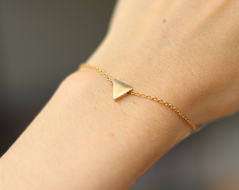 May include: A delicate gold-colored bracelet featuring a small triangular charm. The charm is centered on a fine chain, creating a minimalist and elegant design. The bracelet is shown on a wrist.