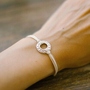 Puede incluir: Una pulsera plateada con un centro circular y cordones color crema. La pulsera se lleva en la muñeca, mostrando el diseño y la textura de la joya. El centro de la pulsera tiene un patrón detallado.