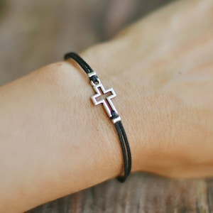 May include: A black cord bracelet with a silver cross charm. The cross is open in the center and has a simple design.