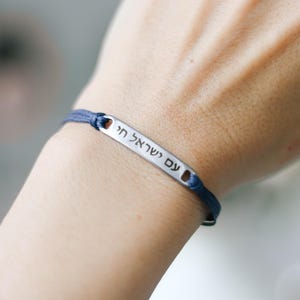May include: A blue cord bracelet with a silver rectangular charm. The charm has Hebrew text engraved on it. The bracelet is worn on a wrist.