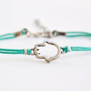 Pu&ograve; includere: Un braccialetto con cordino turchese con un ciondolo a forma di mano di Hamsa color argento. Il braccialetto ha un design delicato con chiusura a nodo scorrevole e dettagli metallici. La mano di Hamsa &egrave; un simbolo di protezione e buona fortuna.