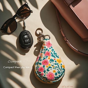 May include: A teardrop-shaped fabric pouch with a floral pattern of pink and yellow flowers on a white background, featuring a metal keyring. Nearby are black sunglasses, a black car key fob, and a pink handbag. Text reads Compact Everyday Size.
