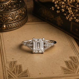 May include: A silver-toned ring featuring a large emerald-cut center stone held by four prongs. The band is detailed with intricate metalwork and small round stones set along the shoulders, extending from the central setting.