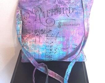 May include: A square crossbody bag with a zip closure and a long strap. The bag features a vintage-style print in shades of purple, blue, and pink, with script and the text "RIFFARD Succursales". The bag is sitting on a black surface.