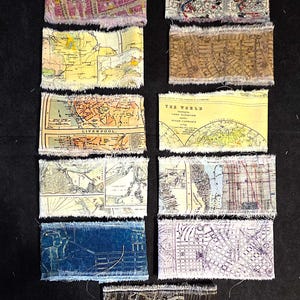 May include: A selection of rectangular fabric pieces, each printed with a vintage map design. The maps depict different cities and regions, in colours such as pink, yellow, blue, and brown. The edges of the fabric are frayed, giving a rustic appearance.
