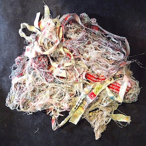 May include: A tangled assortment of fabric scraps, ribbons, and threads in various colors, including red, white, and plaid patterns. Some ribbons have text that reads "PEACE BY", "LET IT SK", and "LIVE IN THE MAGIC + FES".