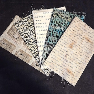 May include: Five rectangular fabric swatches featuring various typographic designs. One has a beige map-like print, one has black letters on white, one has black words on white, one has black letters on a blue-green background, and one has black text.