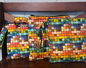 May include: Three square fabric pouches featuring a vibrant mosaic pattern of small, multicoloured squares in shades of orange, yellow, blue, green, and brown. Each pouch includes a matching fabric wrist strap and a top zip closure for storage.