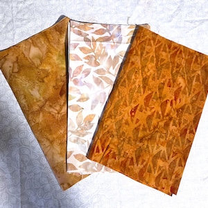 May include: Three rectangular fabric swatches arranged in a fan shape. The left swatch is a mottled golden brown, the center swatch features a white background with light brown leaf patterns, and the right swatch is a deep orange with dark brown geometric shapes.