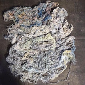 Peut inclure: Un amas emmêlé de fils fins et fibreux dans une palette de couleurs sourdes composée de bleu pâle, de jaune doux, de gris clair et de blanc cassé. Les fibres sont nouées et entrelacées de manière complexe, formant une forme organique dense.