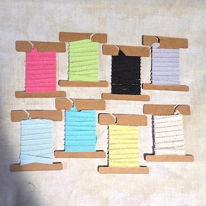 Pode incluir: Vista aérea de oito carretéis de fita colorida. Os carretéis de cartão contêm fitas nas cores rosa, verde, preto, lavanda, azul claro, turquesa, amarelo e branco. As fitas são texturizadas.