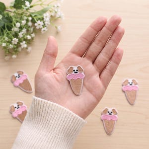 May include: Embroidered iron-on patches featuring a tan dog head emerging from a pink ice cream scoop inside a tan waffle cone. The patches are small, detailed, and shaped like a dessert treat with a canine face on top.