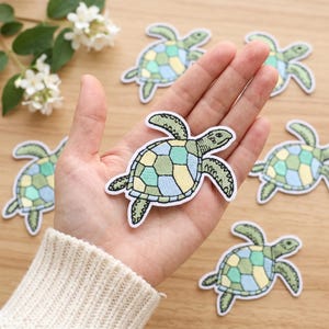 Puede incluir: Una colección de parches bordados de tortugas marinas con un patrón geométrico en el caparazón en tonos verde salvia, azul claro y amarillo pálido. Cada parche tiene un borde blanco y costuras detalladas que definen la forma de la tortuga.