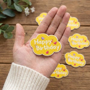 以下が含まれることがあります： 黄色い雲の形をした刺繍ワッペンで、白い文字でHappy Birthdayと書かれ、ピンクと紫の小さなハートがアクセントになっています。ワッペンには白い縁取りがあり、手のひらに乗せられており、近くには同じワッペンがいくつか散らばっています。