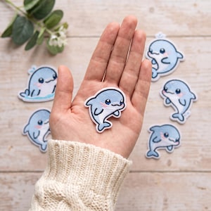 Puede incluir: Una colección de parches bordados termoadhesivos que muestran un delfín azul claro con vientre blanco, ojos azul oscuro y mejillas rosadas. Cada parche tiene un borde blanco y representa al delfín en varias poses de nado estilo dibujo.